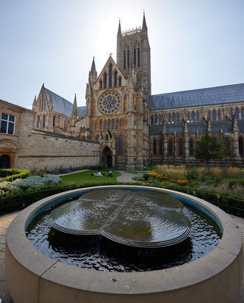 Lincoln cathedral water feature This architectural photograph, taken in the late morning during early autumn, features the water feature and fountain located in the grounds of Lincoln Cathedral in Lincoln, England, United Kingdom. The image prominently displays the historic cathedral, known for its Gothic architecture, with its large west front and iconic central tower rising above the scene. In the foreground, the circular fountain adds an element of stillness to the composition, its surface gently rippling beneath the sunlight. The cathedral, built as a place of Christian worship and an iconic church in England, is a well-known landmark with intricate stonework and the large rose window visible above the main entrance. The garden surrounding the fountain is in late summer bloom, contributing to the seasonal atmosphere of the photograph. The overall scene exemplifies English ecclesiastical architecture, connecting the fountain and landscaping with the historic structure of Lincoln Cathedral in an urban landscape setting.